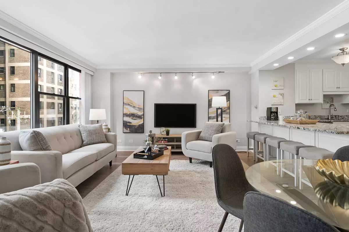 The living room, kitchen open concept at 850 North Dewitt Place Unit 4H in Chicago's Streeterville neighborhood. Listed for sale by The DelGreco Team from Coldwell Banker Realty's Lincoln Park Office. The living room is all white walls and a big panel of windows on the left with the open kitchen on the right. A white couch sits under the window and a TV flanked by vertical abstract art on each side is on the center wall in the middle of the image.