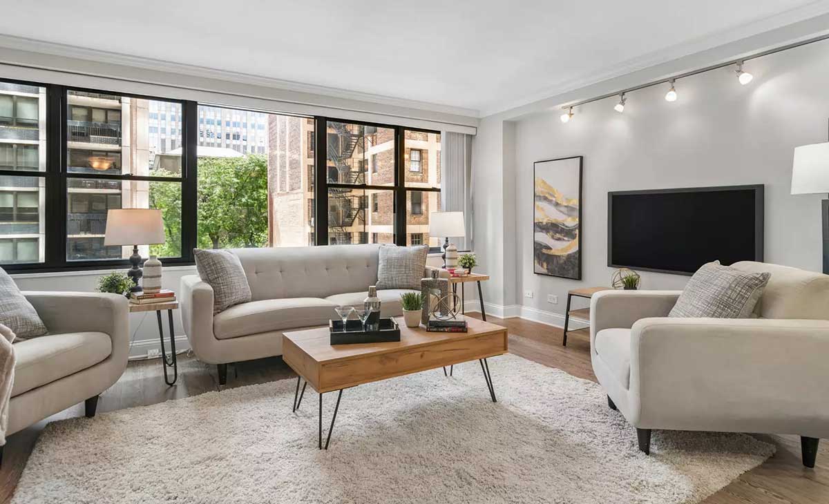 The living room at 850 North Dewitt Place Unit 4H in Chicago's Streeterville neighborhood. Listed for sale by The DelGreco Team from Coldwell Banker Realty's Lincoln Park Office. The living room is all white walls and a big panel of windows on the left and the TV mounted on the wall on the right.