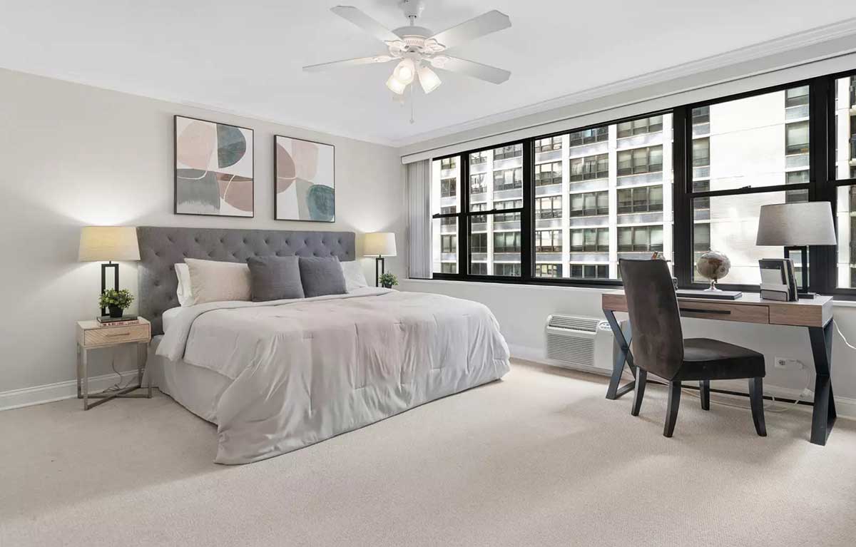 The primary bedroom at 850 North Dewitt Place Unit 4H in Chicago's Streeterville neighborhood. Listed for sale by The DelGreco Team from Coldwell Banker Realty's Lincoln Park Office. The bedroom is all white walls and carpet. The wall on the right is a large spread of big windows. The king-sized bed is in the middle of the image up against the wall on the left.